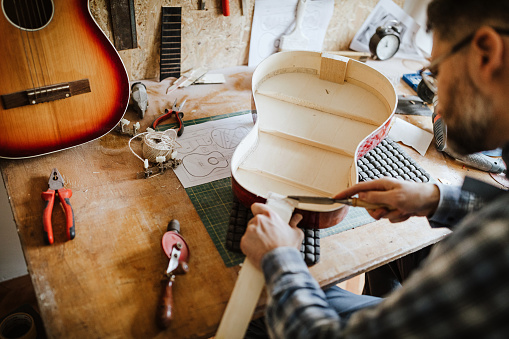 Mid-adult man sitting at workbench and repairing acoustic guitar at his workshop