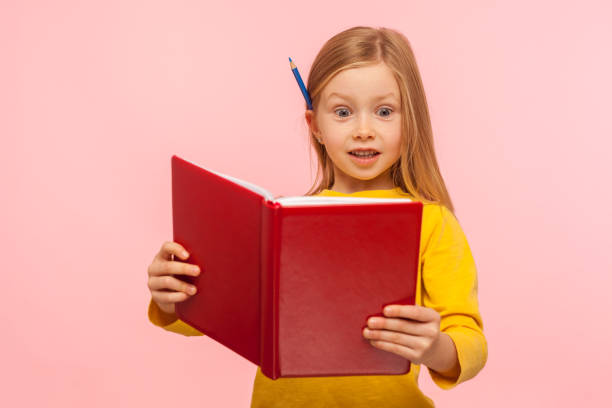 шокирующие факты. удивленная умная маленькая девочка с карандашом за ухом читает большую книгу с удивленным выражением - surprise child little girls shock стоковые фото и изображения