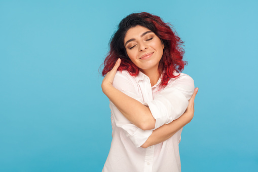 I love myself. Portrait of beautiful narcissistic woman with fancy red hair embracing herself with pleased satisfied face expression, positive self-esteem. studio shot isolated on blue background