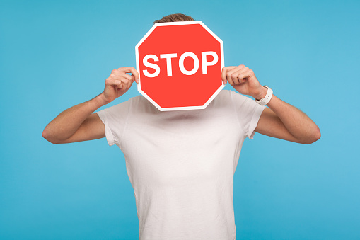 Portrait of unknown man in white t-shirt covering face with Stop symbol, anonymous person holding red traffic sign, warning to go, prohibition concept. indoor studio shot isolated on blue background