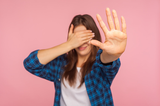 No, don't show me! Portrait of confused girl in checkered shirt covering eyes with hand and showing stop gesture, afraid to look at shameful content. indoor studio shot isolated on pink background