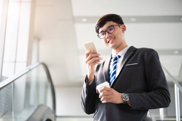 istock A asian handsome young businessman holding hot coffee cup while using his phone and standing on the escalator. Business concept. 1213375808