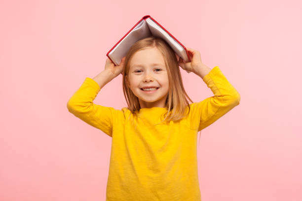 ritratto di felice pigro bambina che copre la testa con libro e sorridente alla macchina fotografica, bambino disobbediente divertirsi - schoolgirl school children isolated child foto e immagini stock