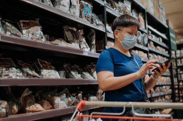 une femelle chinoise asiatique portant un masque sortant faisant des emplettes dans le supermarché regardant la liste de vérification de téléphone - shopping mall shopping sale retail photos et images de collection