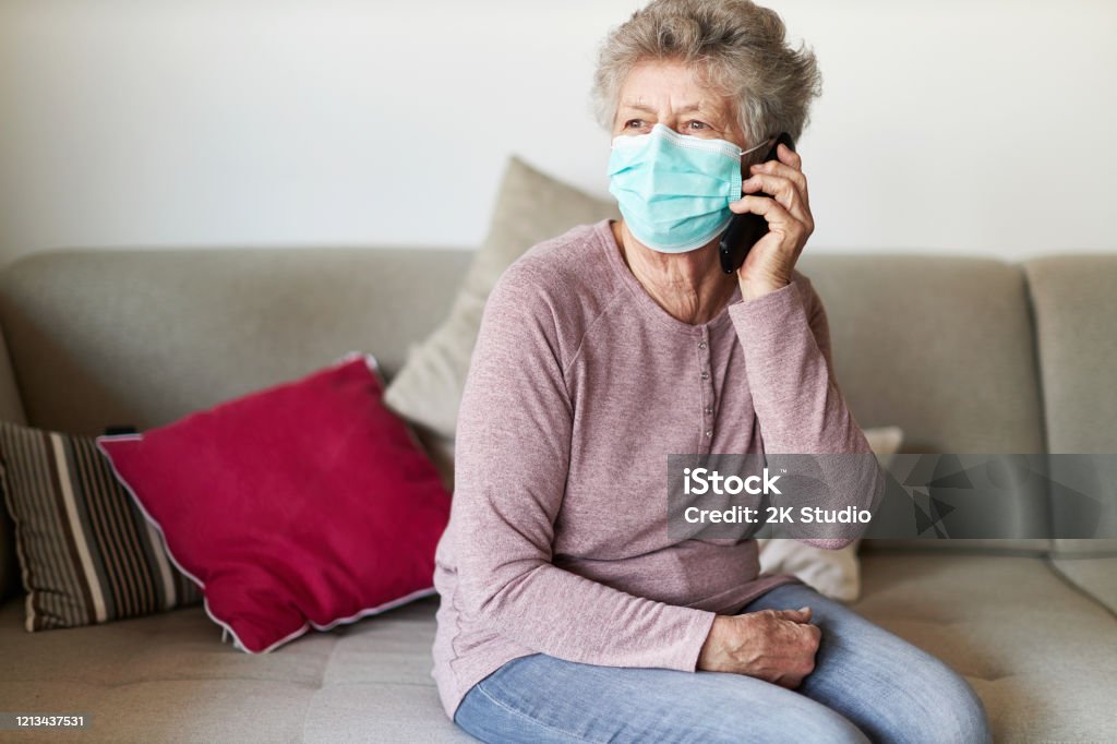 A senior citizen sits alone on her sofa in the living room wearing a respirator or surgical mask A senior citizen sits alone on her sofa in the living room wearing a respirator or surgical mask and looks sadly and frightened out of the window and into the camera while on the phone Senior Adult Stock Photo