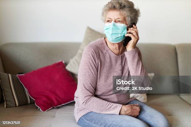 A Senior Citizen Sits Alone On Her Sofa In The Living Room Wearing A Respirator Or Surgical Mask Stock Photo - Download Image Now