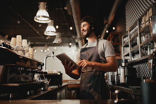 Happy young coffee shop owner holding using digital tablet