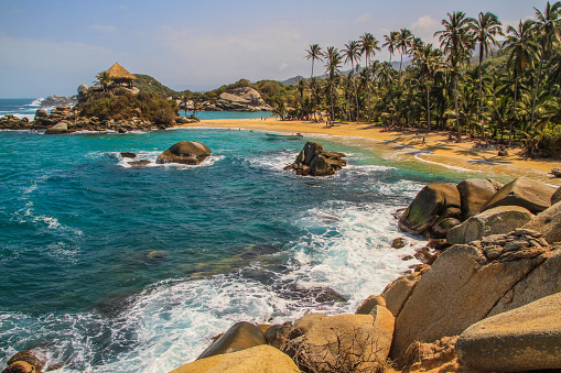 Beautiful tropical beach with famous landmark of a beach hut and blue ocean with white waves, palm trees and rain forest at Tayrona National Park, Colombia