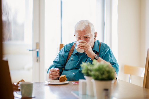 Senior Caucasian man sitting at a table drinking a cup of tea as he finishes his cake.