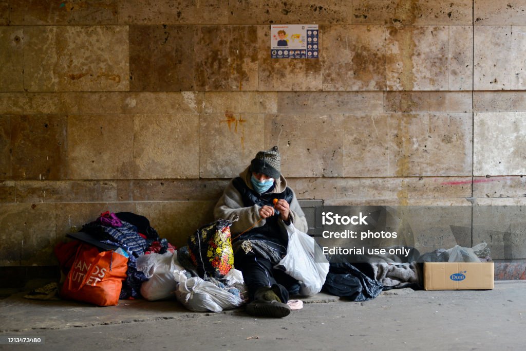 Una mujer sin hogar con máscara médica se sienta en un pasaje subterráneo cerca de un metro cerrado. - Foto de stock de Sin techo libre de derechos