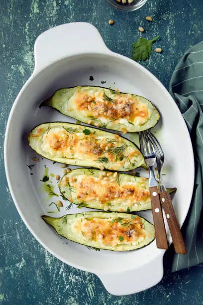 Baked zucchini stuffed with cottage cheese, parmesan, garlic, dill and pine nuts in baking dish. Green shabby rustic background.