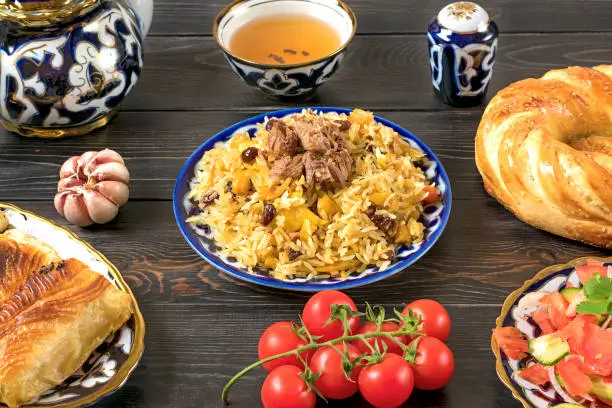 National Uzbek pilaf with meat, achichuk salad of tomato, cucumber, onion in plate with traditional pattern, cilantro, cherry tomatoes, garlicbread tortilla - patir on dark wooden table Top view.