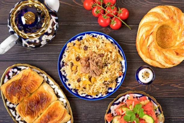 National Uzbek pilaf with meat, achichuk salad of tomato, cucumber, onion in plate with traditional pattern, cilantro, cherry tomatoes, garlicbread tortilla - patir on dark wooden table Top view.