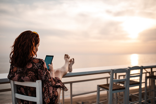 Cheerful young woman relaxing at the beach coffee shop, surfing the net on her tablet while enjoying the view of the Ionian sea