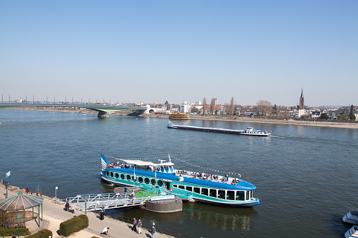 River Rhine and tourboat at promenade in springtime seen from higher angle. People are on promenade and on boat. A freight ship is passing Rhine upstream