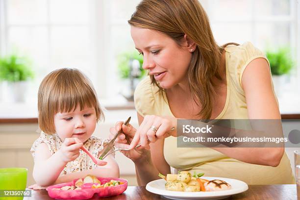 Pregnant Mother In Kitchen Eating Meal Helping Daughter Eat Stock Photo - Download Image Now