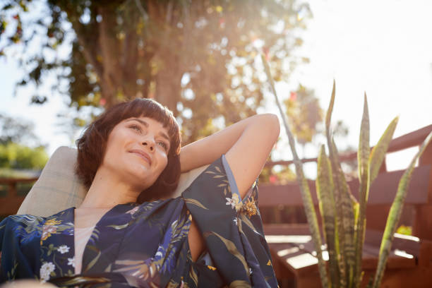 Smiling young woman lying back in a patio deck chair Young woman looking deep in thought and smiling while lying back in a deck chair on her patio on a sunny afternoon porch photos stock pictures, royalty-free photos & images