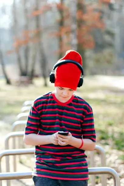 March 18, 2020 - Kabaty, Poland: cute stylish Caucasian boy using his phone while wearing red hat, striped shirt, headphones, being concentrated.
