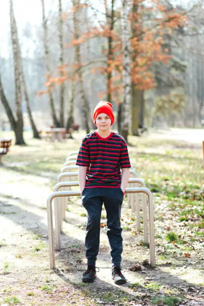 March 18, 2020 - Kabaty, Poland: smiling cute teen (11-12) boy in red hat, striped colourful t-shirt leaning on bike rack enjoying early spring in beautiful park