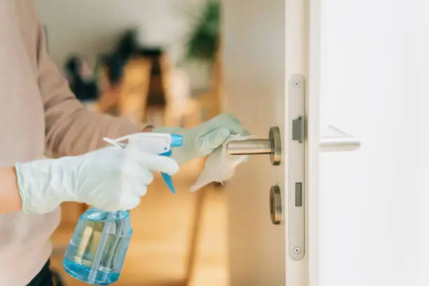 Woman in disposable gloves cleaning a door handle with a disinfection spray