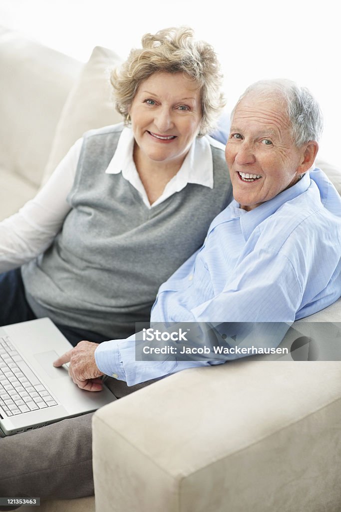 Coppia di anziani guardando la telecamera mentre lavorando su computer portatile - Foto stock royalty-free di Abbigliamento casual