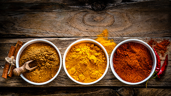 Ground scented spices arranged in a row shot from above on rustic wooden table. The ground turmeric, cinnamon and paprika are in white bowls placed at the bottom of an horizontal frame leaving useful copy space for text and/or logo at the top. High resolution 42Mp studio digital capture taken with Sony A7rII and Sony FE 90mm f2.8 macro G OSS lens