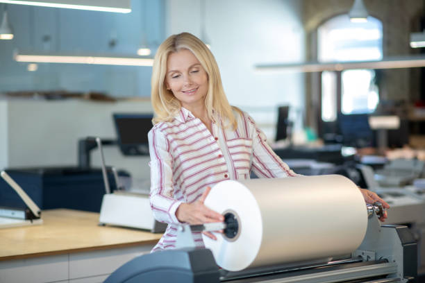 istock Blonde woman standing at paper roll machine, checking the roll 1213557012