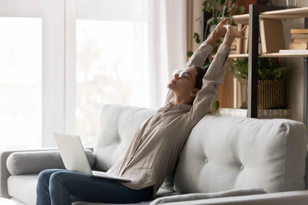 Happy woman stretching sit on comfortable couch with laptop Happy young woman stretching sit on comfortable couch with laptop, lazy girl relaxing doing exercise take break after using computer rest on sofa enjoy stress free day feeling muscle relief at home meditation room office stock pictures, royalty-free photos & images