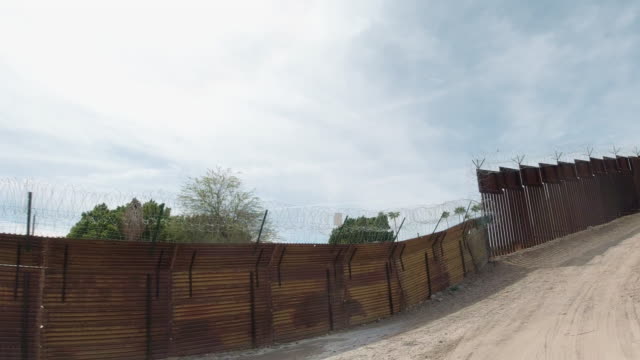 Dolly Shot of a Dirt Road Running Parallel to Parts of the Old Wooden and New Steel-Slat Border Wall Topped by Razor Wire (on the US Side) between Mexico and the United States with the Town of Los Algodones, Mexico on the Other Side on a Partly Cloudy Day
