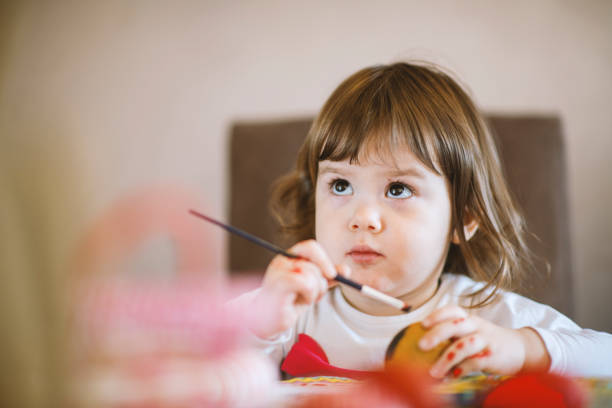 Curious baby girl sitting at table playing with hand colored Easter eggs