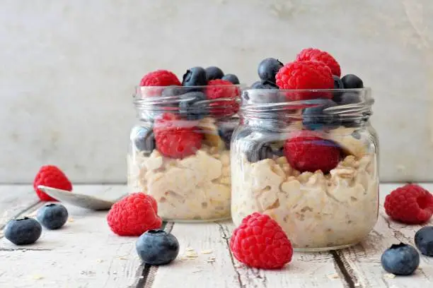 Overnight oats with fresh blueberries and raspberries in jars on a rustic white wood background
