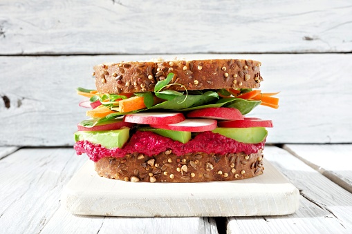 Superfood sandwich with beet hummus, avocado, vegetables and greens on whole grain bread against a white wood background