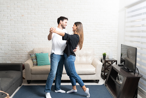 Full length of smiling Latin boyfriend and girlfriend dancing together in living room at home