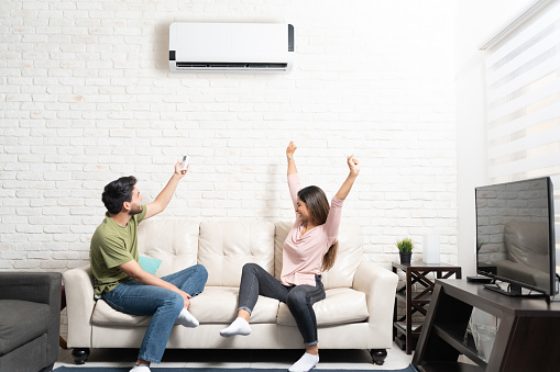 Boyfriend turning on air conditioner while cheerful girlfriend sitting with arms raised on sofa at home