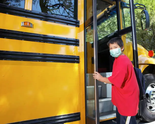 Young elementary school boy climbs on to school bus wearing surgical mask