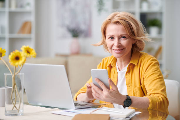 sorridente donna d'affari bionda matura con smartphone che ti guarda - furniture internet adult blond hair foto e immagini stock