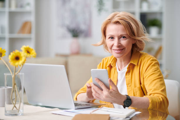 smiling mature blond businesswoman with smartphone looking at you - senior adult happiness computer looking at camera imagens e fotografias de stock