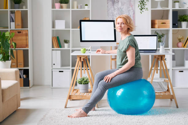 donna matura attiva seduta su fitball da scrivania davanti al monitor del computer - yoga business women living room foto e immagini stock