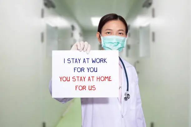 Asian doctor at hospital wear medical masks, holding paper with text I STAY AT WORK FOR YOU, YOU STAY AT HOME FOR US. stay at home policy campaign to control COVID-19 Coronavirus outbreak situation