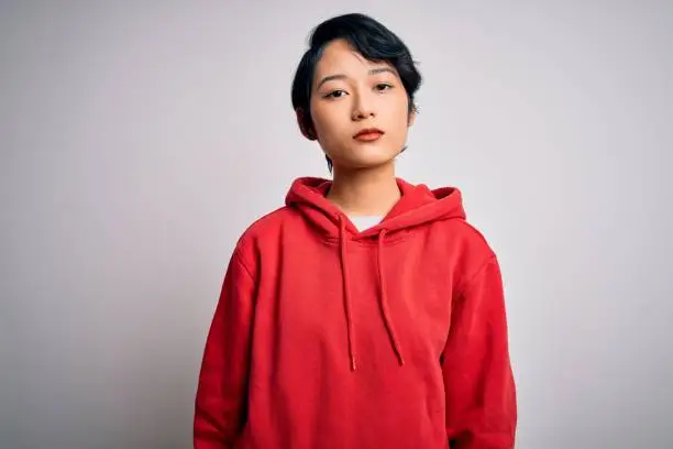 Photo of Young beautiful asian girl wearing casual sweatshirt with hoodie over white background Relaxed with serious expression on face. Simple and natural looking at the camera.