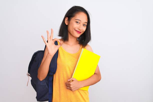 istock Young chinese student woman wearing backpack holding book over isolated white background doing ok sign with fingers, excellent symbol 1213661774