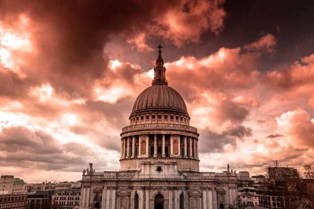 Color image depicting the famous dome and ancient architecture of St Paul's cathedral in London, UK. The cathedral is surrounded by a glowing orange sunset and cloudscape.