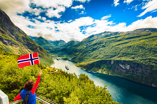 Female tourist with norwegian flag enjoying scenic view over fjord Geirangerfjord with cruise ship. Cruising vacation and travel.