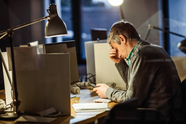 tired businessman using mobile phone while working late in the office. - looking through window businessman problems reading imagens e fotografias de stock