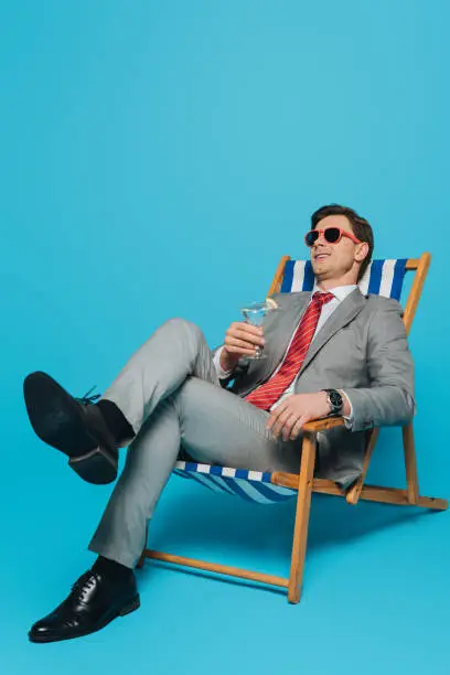 Photo of happy businessman in sunglasses sitting in deck chair with glass of cocktail on blue background