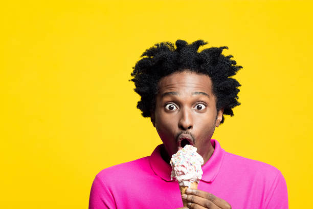 Surprised young man eating gelato Portrait of shocked afro american young man wearing pink polo shirt, holding big ice cream in hands, staring at camera, rolling eyes. Studio shot on yellow background. licking ice cream cone stock pictures, royalty-free photos & images