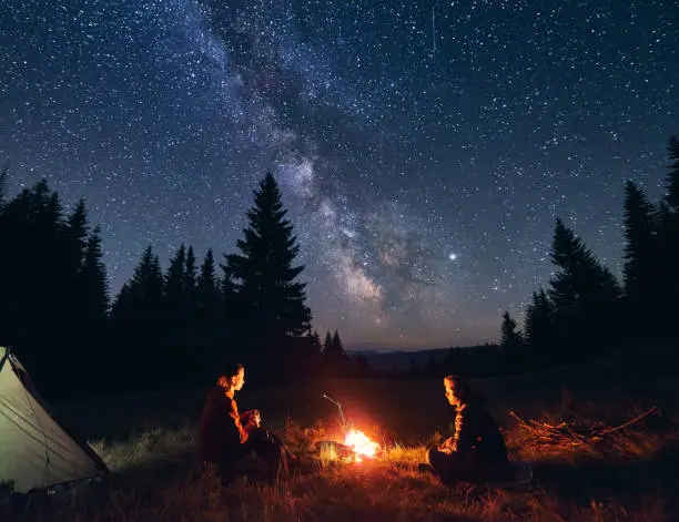 Photo of Man and woman communicate by fire against background of starry sky with bright Milky way. Night camping.