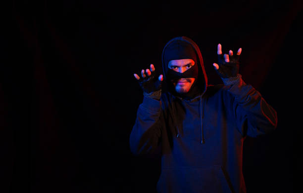 istock Robber, a thug in a balaclava raised his hands on a black background, surrenders. Robbery, hacker, crime, theft, arrest. Copy space 1213691606