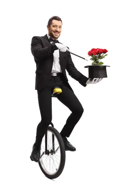 Full length portrait of a smiling magician iding a unicycle and holding wand and hat with flowers isolated on white background