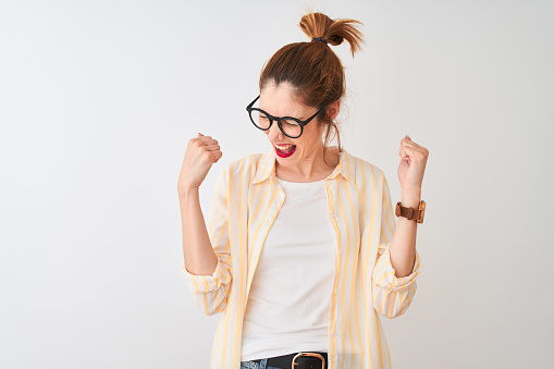 Redhead woman wearing striped shirt and glasses standing over isolated white background very happy and excited doing winner gesture with arms raised, smiling and screaming for success. Celebration concept.